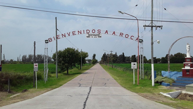 Entrada rural con un arco metálico que dice "Bienvenidos a A. Roca" sobre un camino pavimentado rodeado de campos verdes y un monumento a la derecha.

Description generated by AI